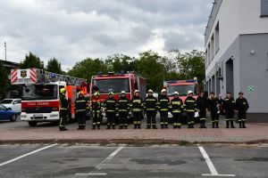 Zdjęcie: Strażacy w mundurach na tle wozów strażackich i remizy